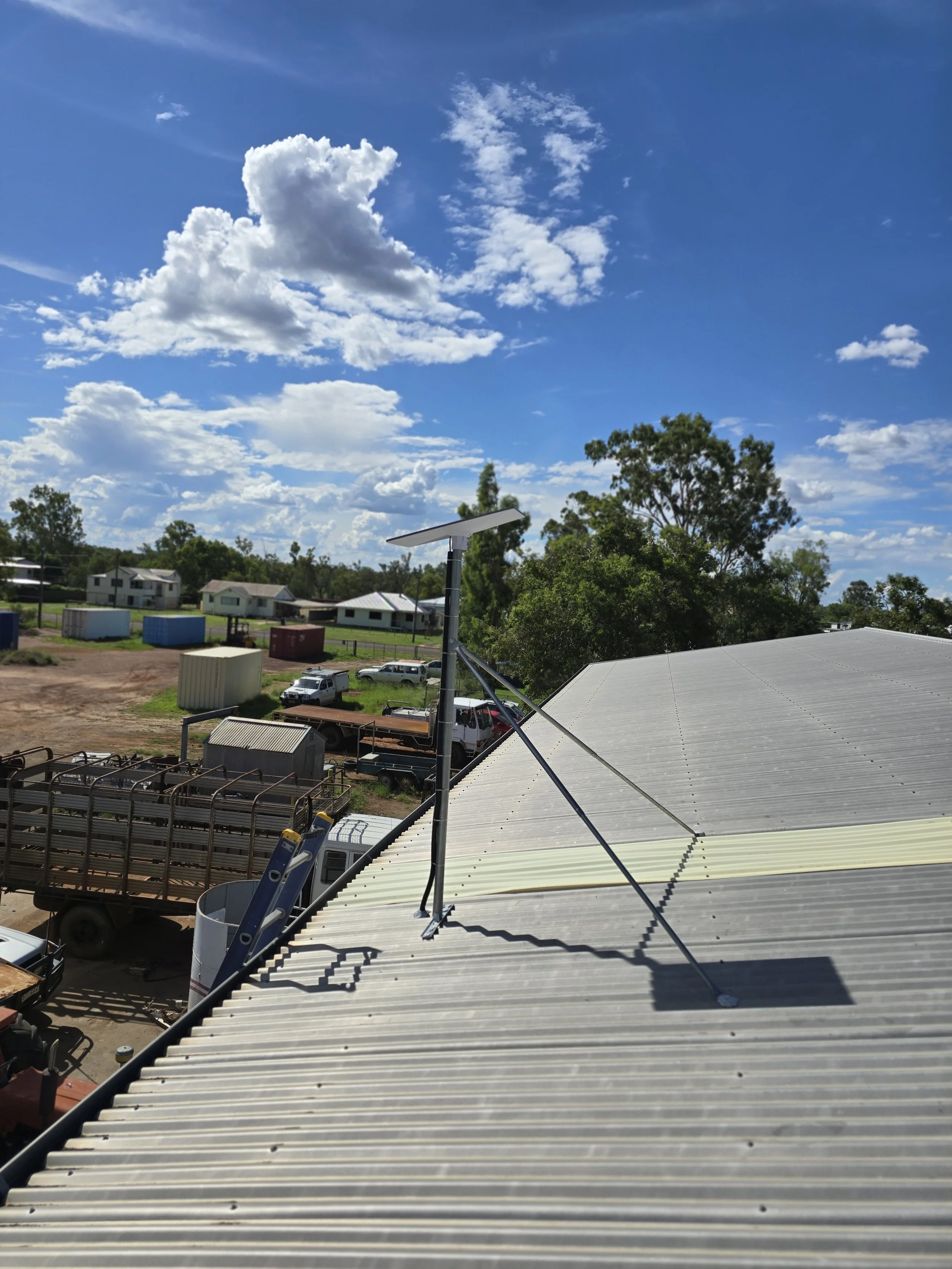 View from a building rooftop showing a solar panel, vehicles, storage containers, trees, residential houses, and a blue sky with scattered clouds.