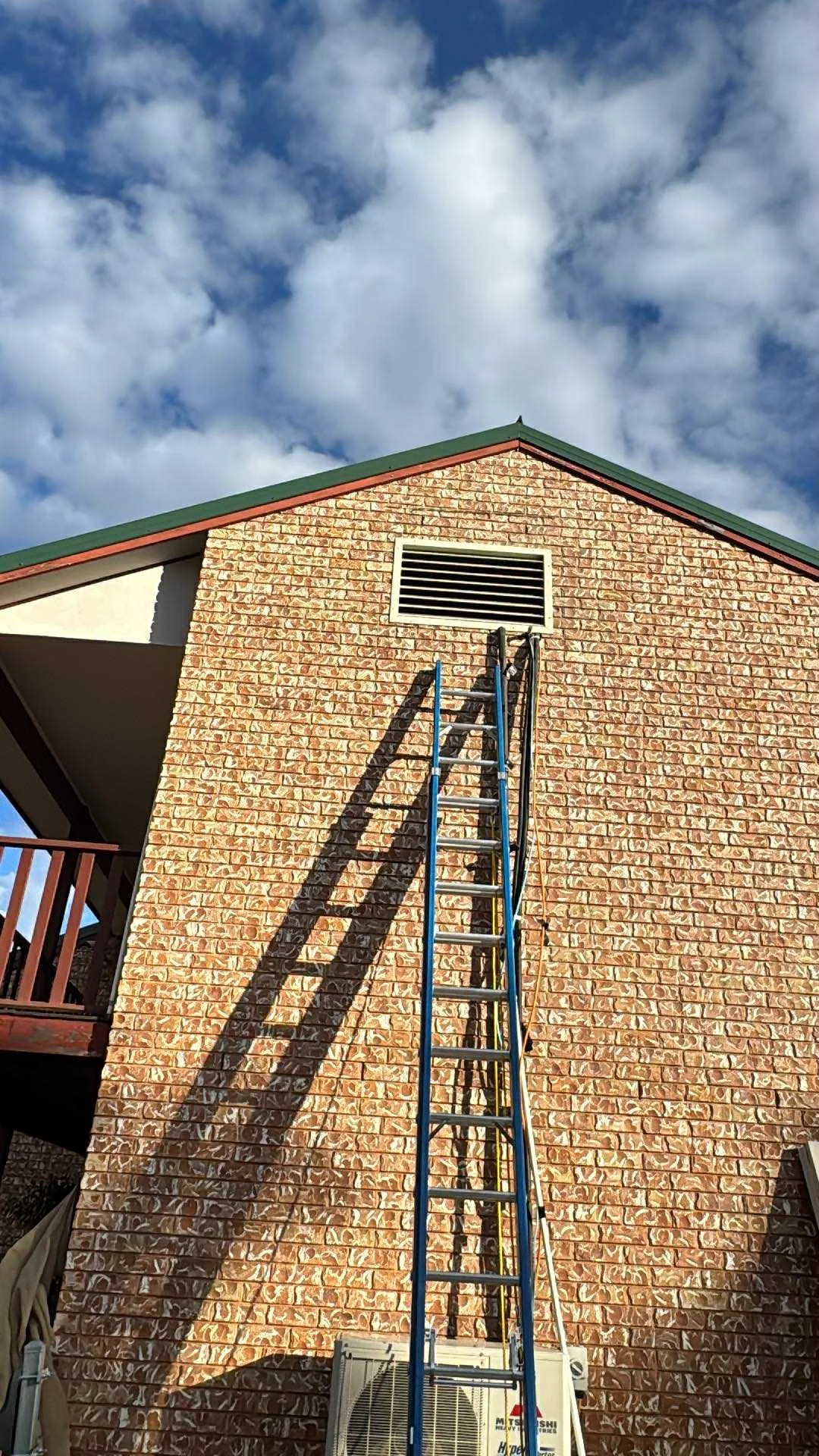 A brick house with a green roof, a blue ladder leaning against it, a window vent near the roof, and an air conditioning unit at the bottom. The sky is partly cloudy.