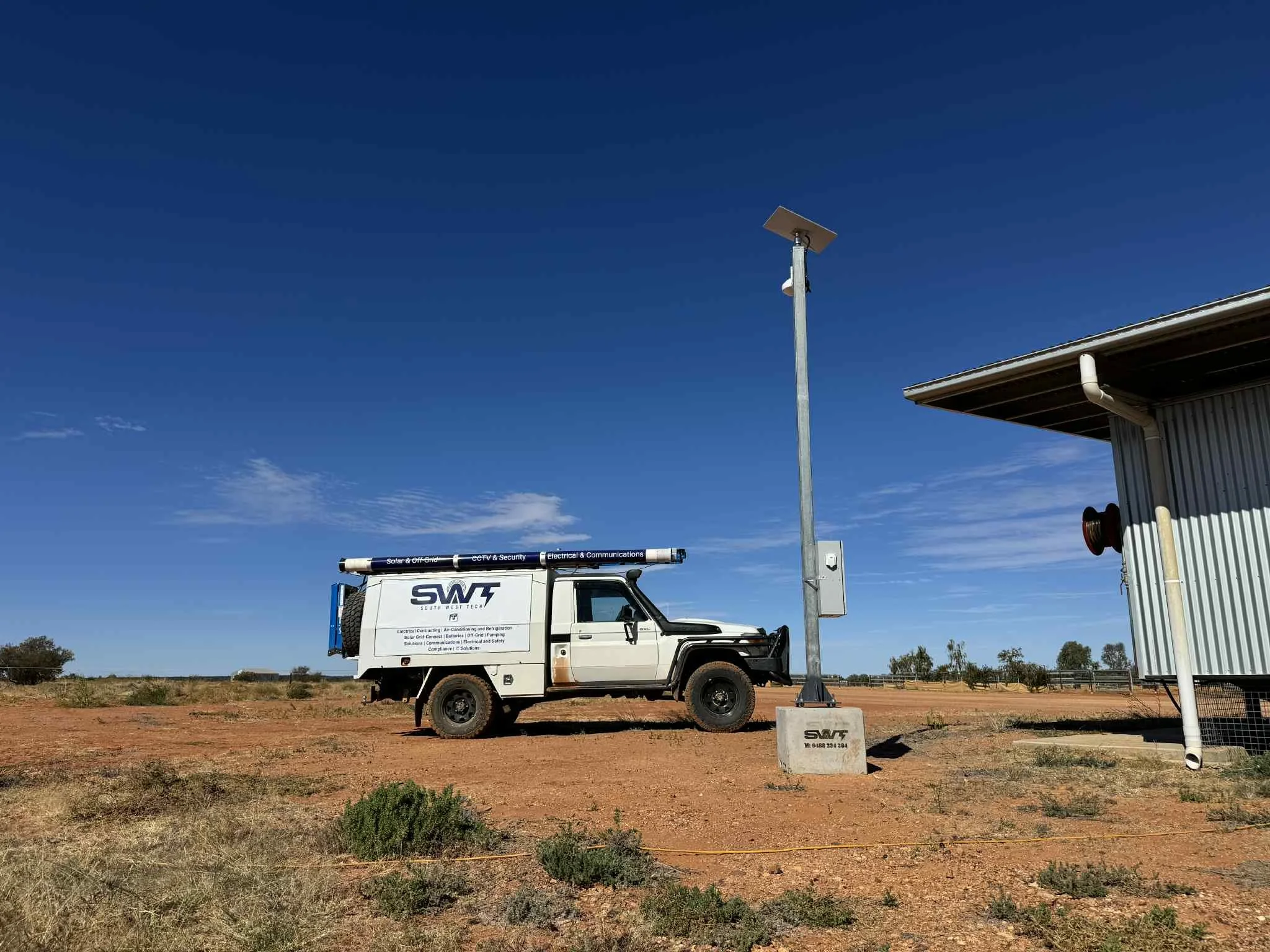 A utility truck parked outdoors on dirt ground next to a building with a solar panel and communication equipment mounted on a tall pole, against a clear blue sky.