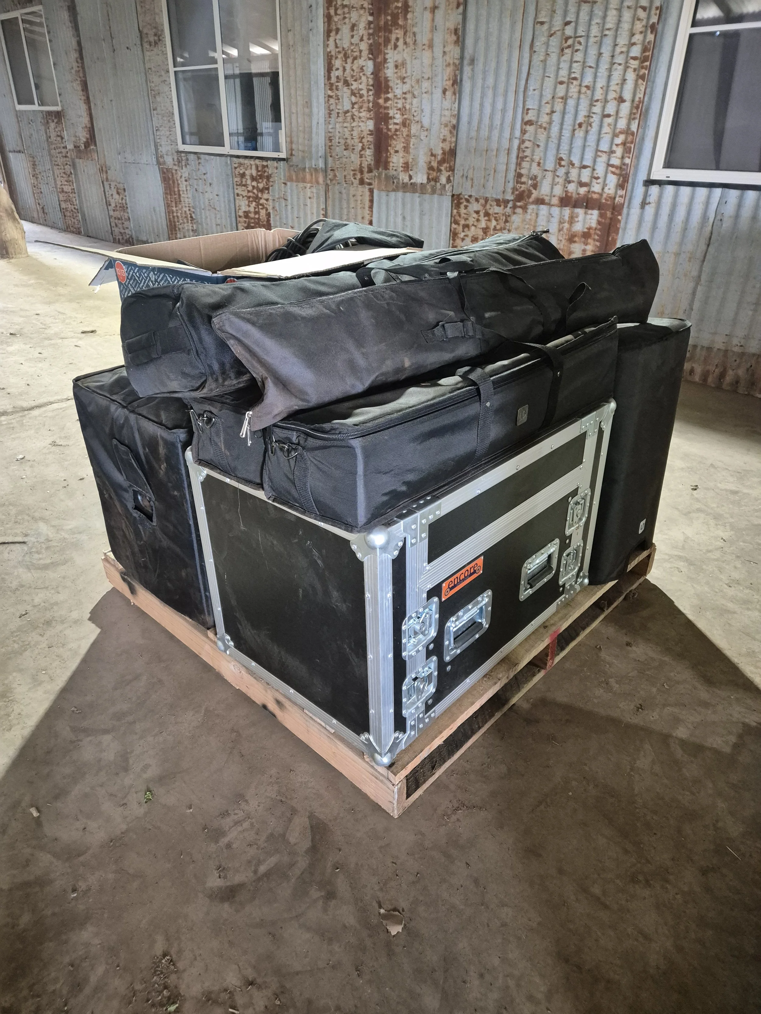 A large black equipment case with metal edges and latches, stacked on a wooden pallet on a concrete floor. On top of the case, there are black bags, folders, and cables. In the background, there is a corrugated metal wall with windows.