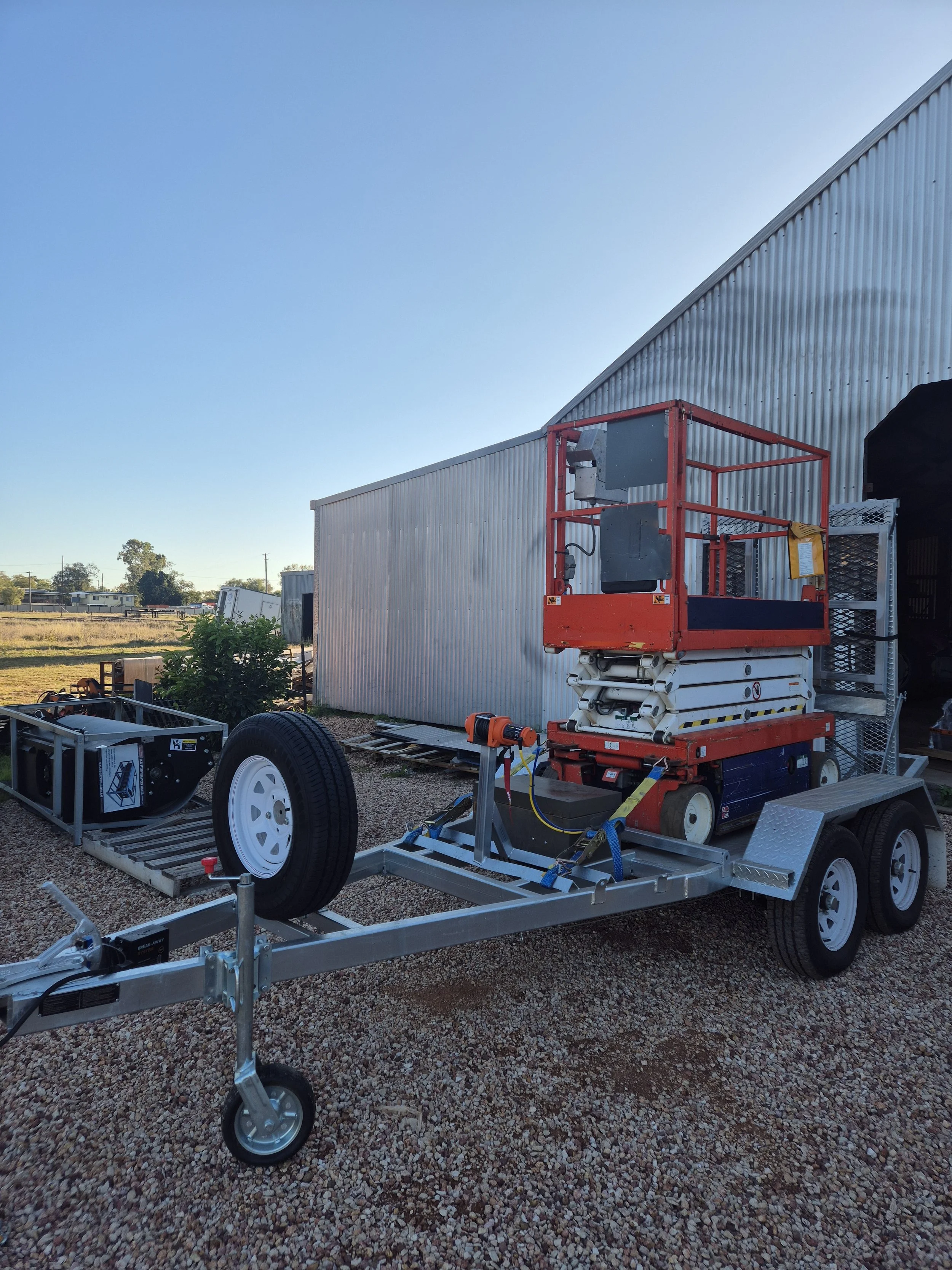 A small trailer with construction equipment and a scissor lift parked on gravel outside a metal building with a curved roof.