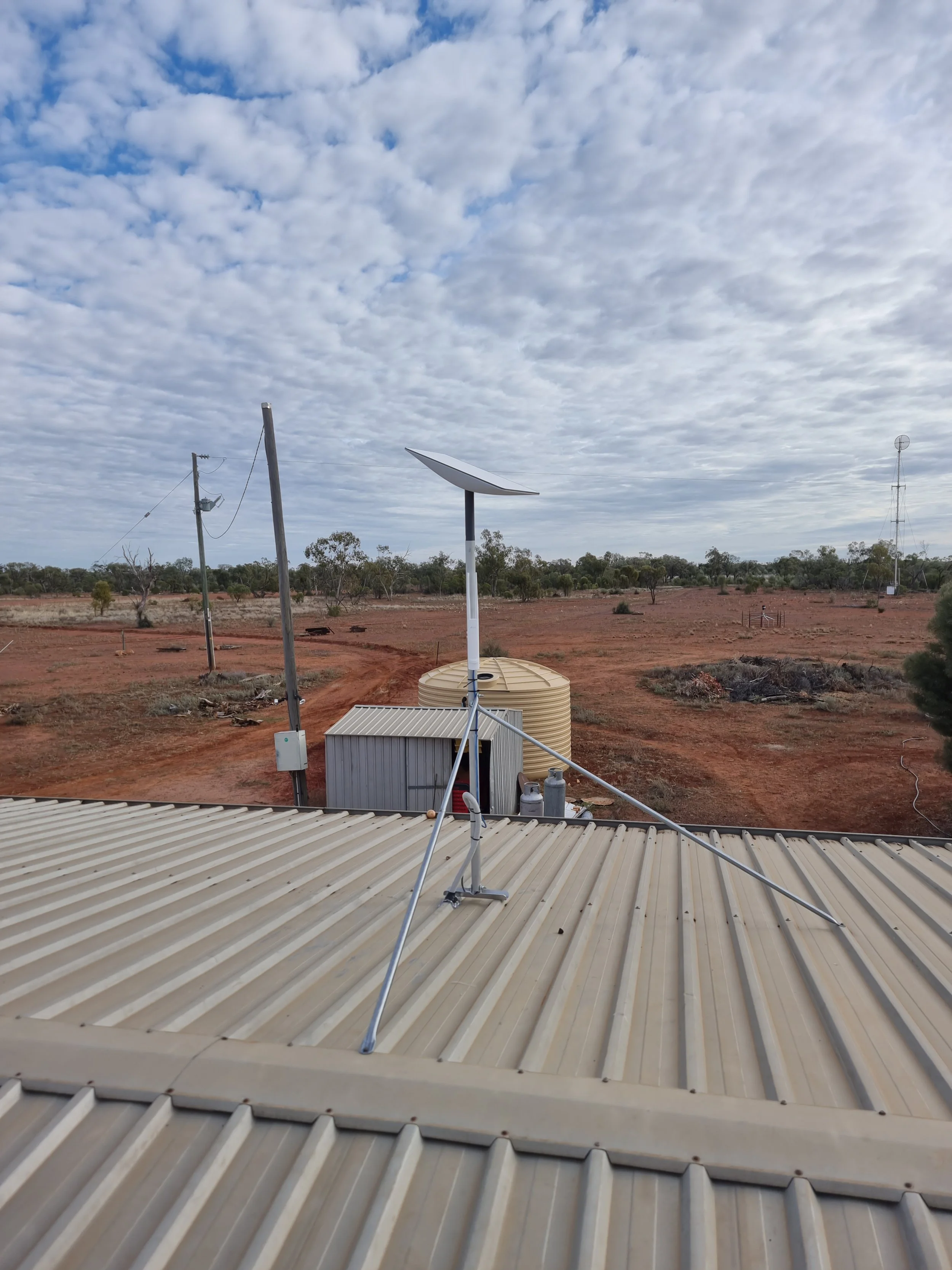View of a rooftop with satellite dishes and antennas in a rural area with a dirt road, sparse trees, and a cloudy sky.