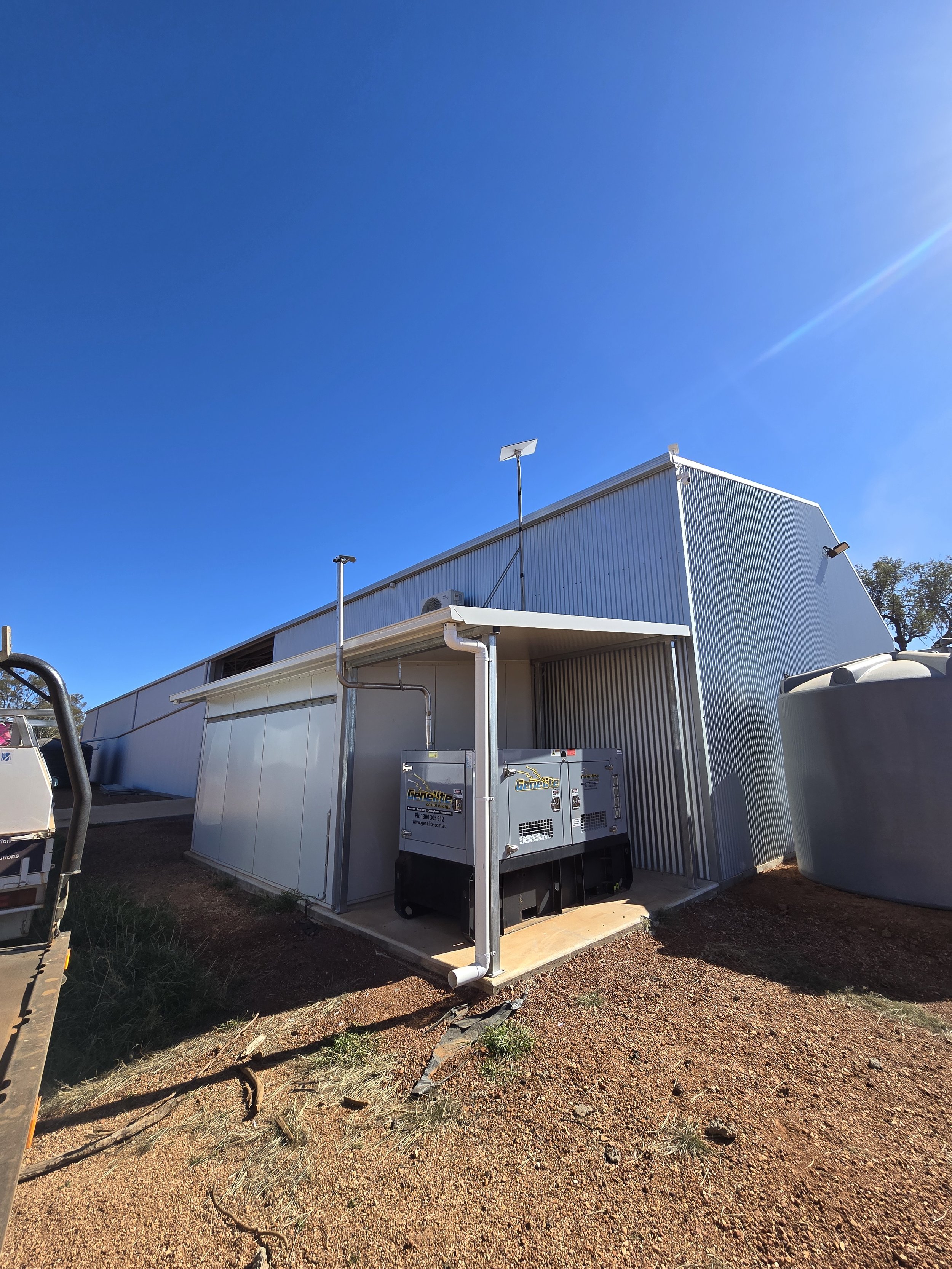 Large industrial building with metal siding, several vents, and a chain-link fence. Equipped with security cameras, solar panels, and utility boxes, under a clear blue sky.