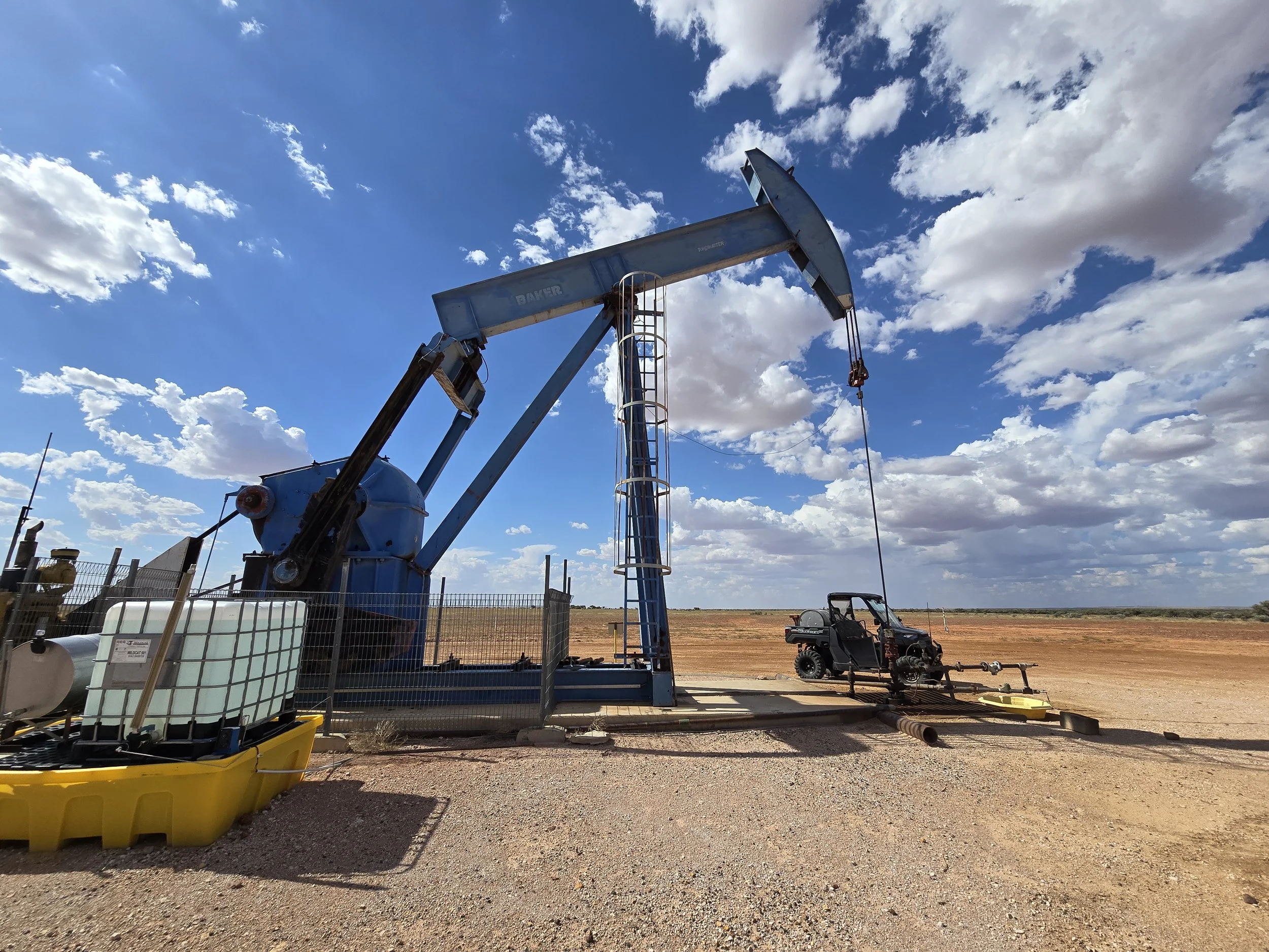 An oil pumpjack operating in a rural area with a clear blue sky and scattered clouds in the background. There is equipment and containers in the foreground and a utility vehicle nearby.
