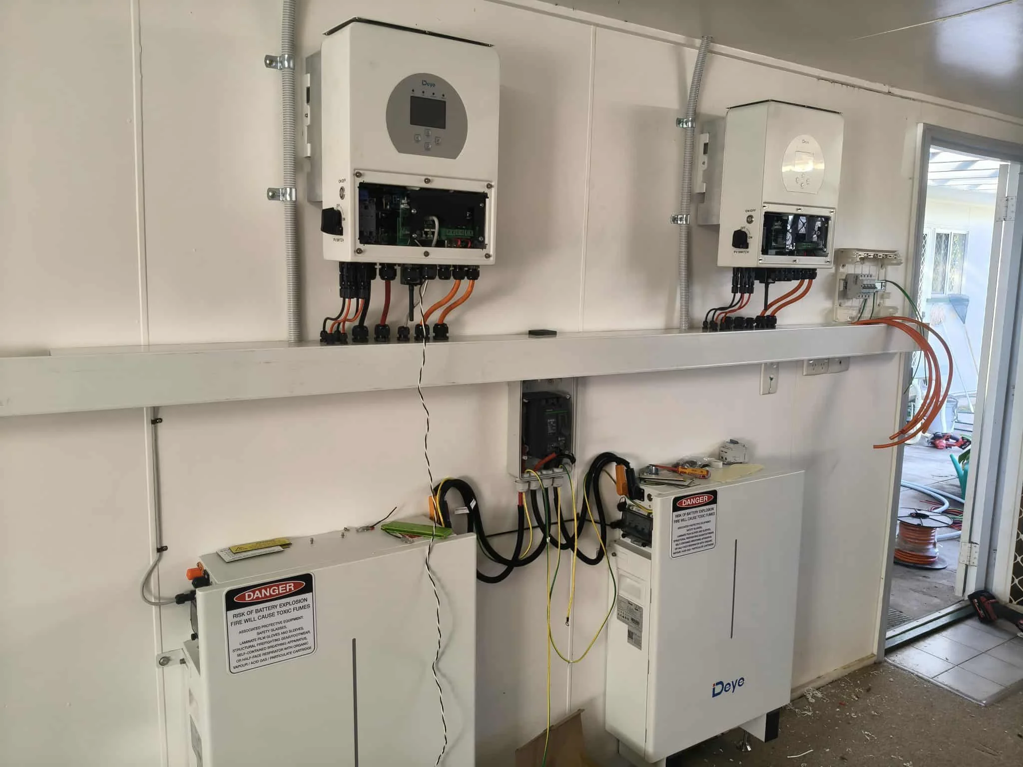 Two large white electrical boxes with open doors mounted on a wall, with visible wiring and safety labels, situated inside a room with a window and exposed flooring.