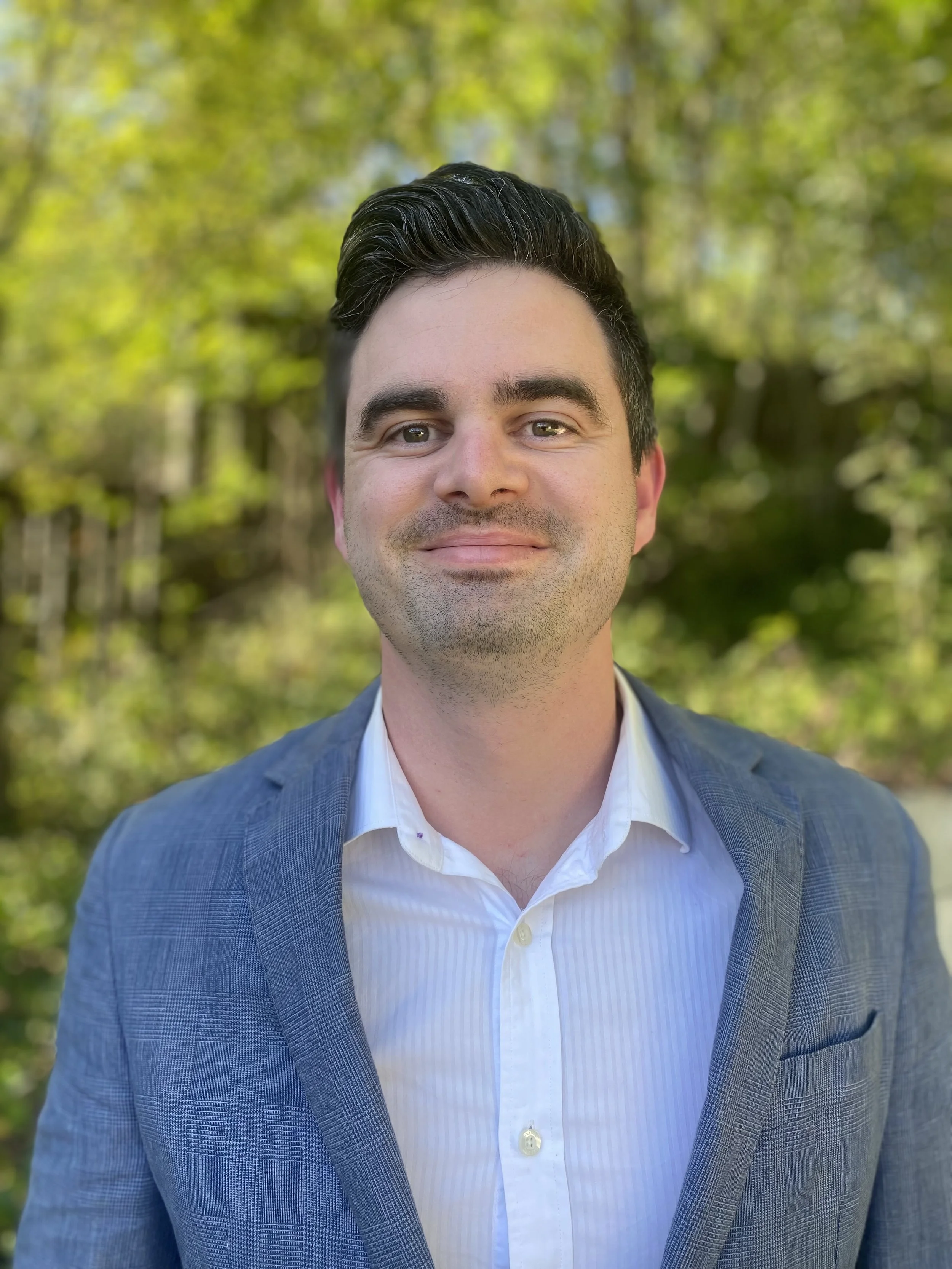 A man with dark, slicked-back hair wearing a light blue suit and white shirt, smiling outdoors with a green, blurred forest background.