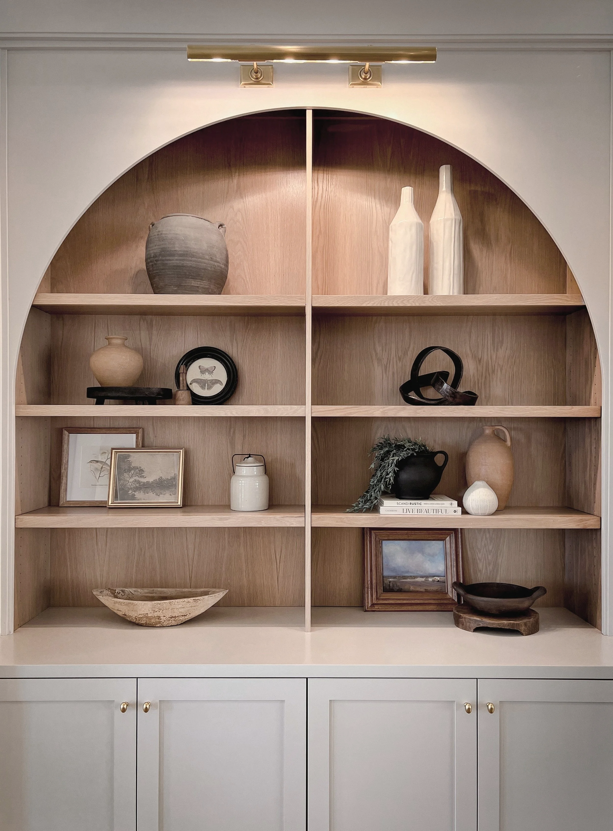 Wooden display cabinet with decorative vases, framed artwork, books, and bowls, topped with a brass light fixture.