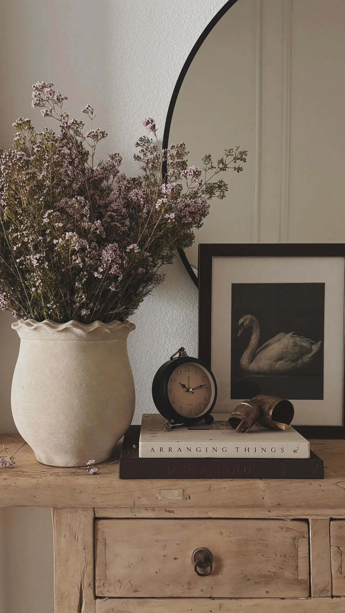 Decorative arrangement featuring a large white ceramic vase with purple flowers, a framed swan picture, a small clock, and vintage objects on a wooden surface, with a mirror in the background.