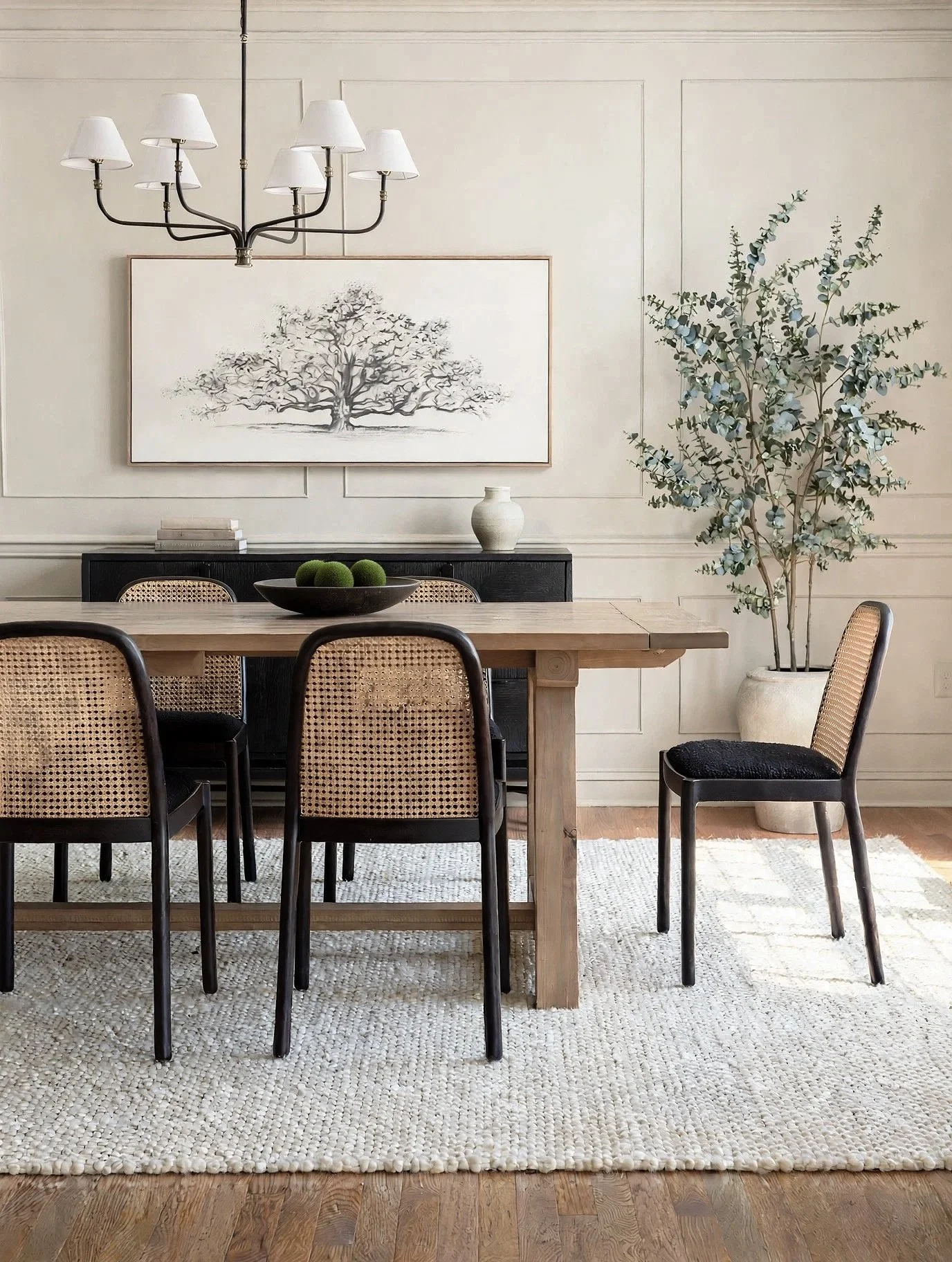 A dining room with a light-colored wall, a large black and white framed art piece of a tree, a wooden dining table with black and woven chairs, a black console with books and a white vase, a large potted eucalyptus plant, and a chandelier with white 