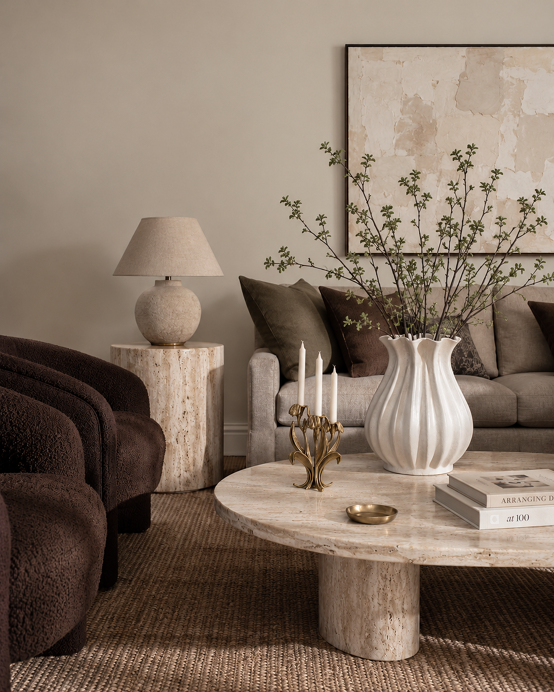 Elegant living room with beige walls, a beige sofa with dark brown and taupe pillows, a marble coffee table with a large white vase holding branches, a gold candelabrum with white candles, and a stack of books. A side table with a textured beige lamp and artwork on the wall.