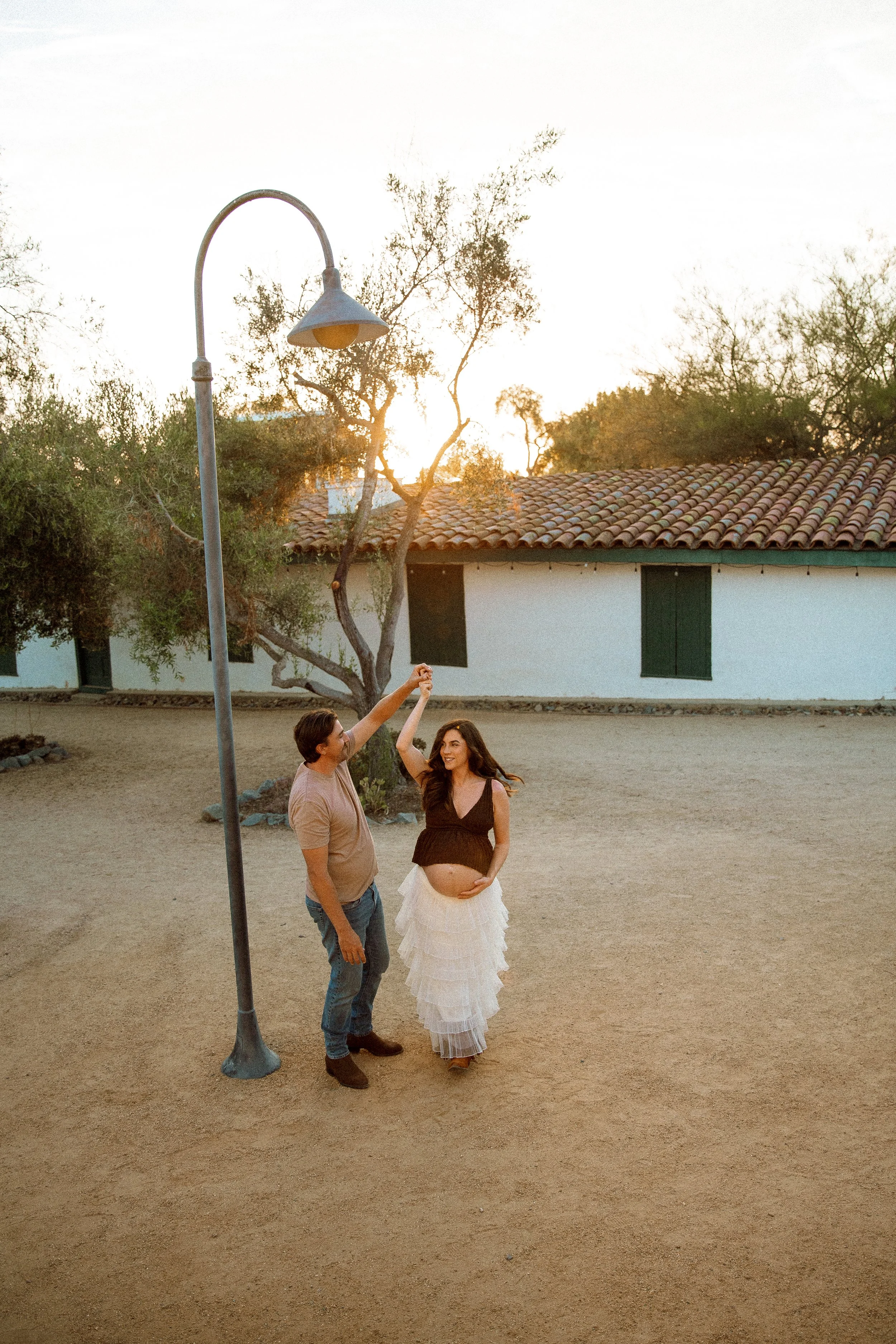 A young woman in a white layered dress with a bare belly and long hair poses with a man next to a vintage streetlamp at sunset. The woman is raising her hand to high five the man, both smiling. The background features trees and a building with a tiled roof.