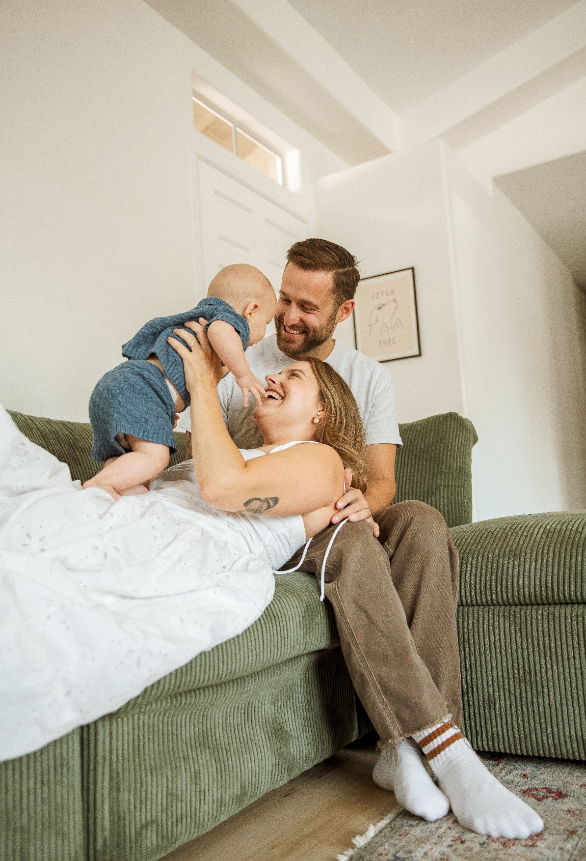 A happy family of three, including a woman, a man, and a small child, enjoying a playful moment on a green couch in a cozy living room with white walls and artwork. The woman is lying on the couch, holding the child up, while the man sits beside them, smiling.