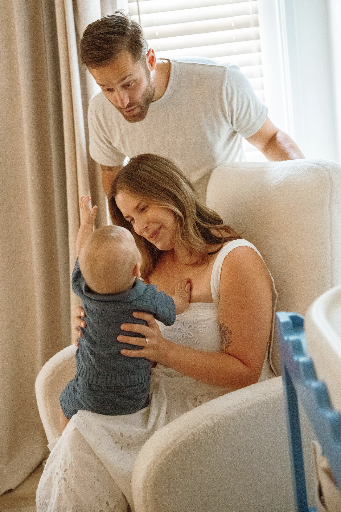 A family scene with a newborn baby, mother, and father; the mother is sitting, breastfeeding the baby with a smile, the father looks at them with interest, all in a cozy room near a window.