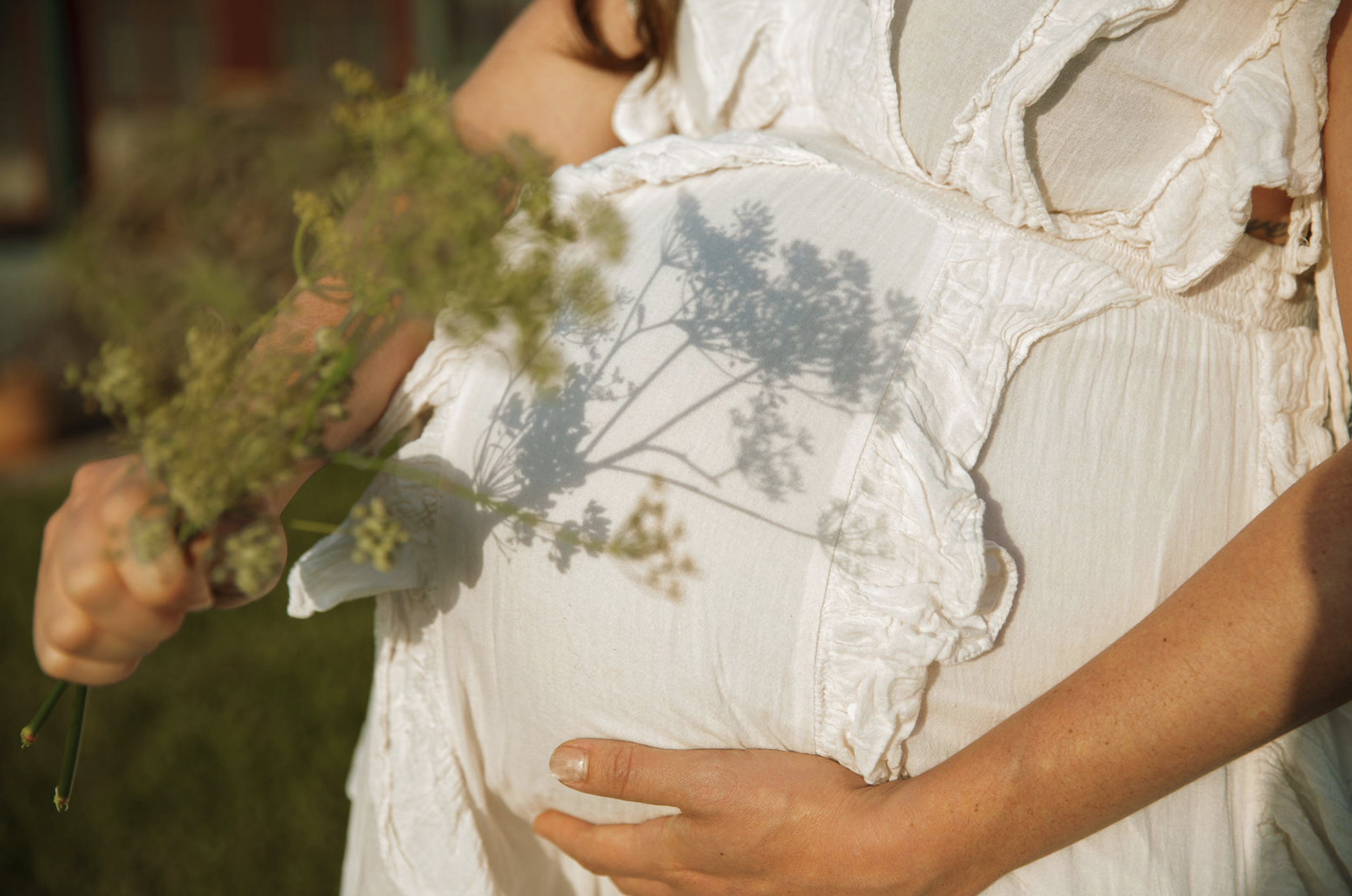 A soon-to-be mother wearing a white, textured dress holding a small bouquet of green flowers outdoors, with their hand resting on their stomach and the shadow of a tree cast on their dress.