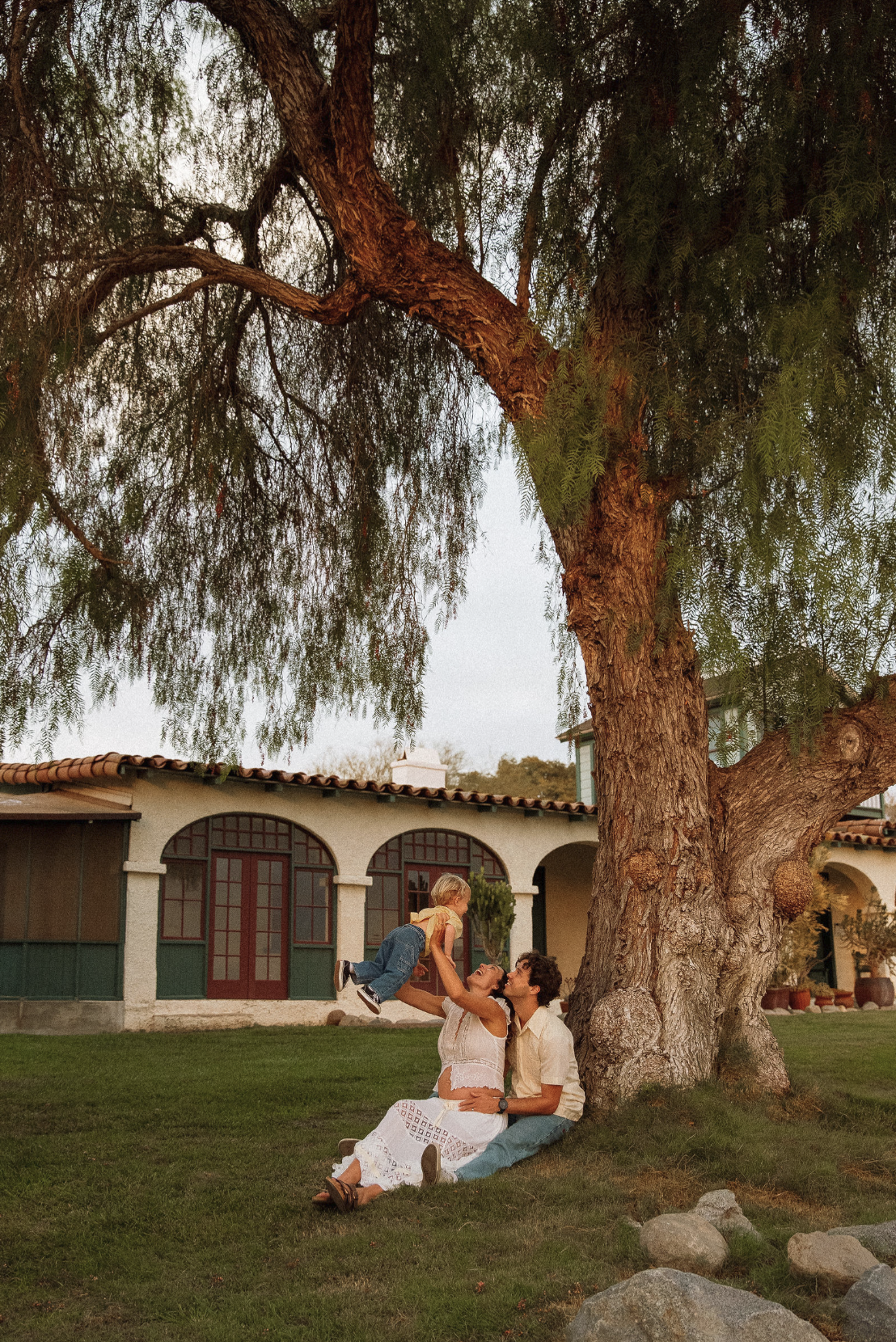 A family of three enjoying a moment under a large tree in front of a house during sunset. The father, mother, and young child are playing and smiling, with the child being lifted into the air by the parents.