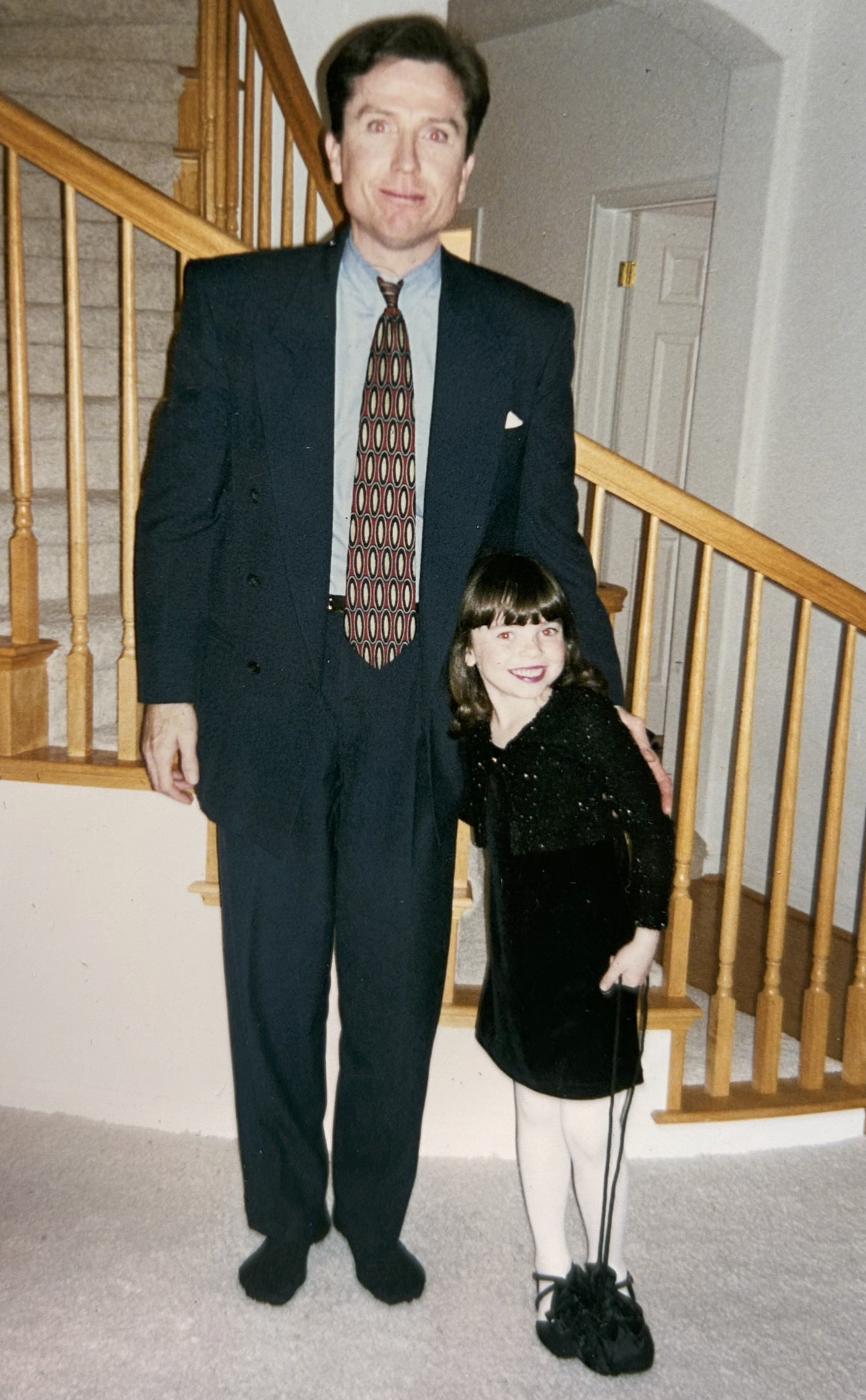 A man in a suit standing next to a young girl, both smiling, inside a house with a staircase in the background.