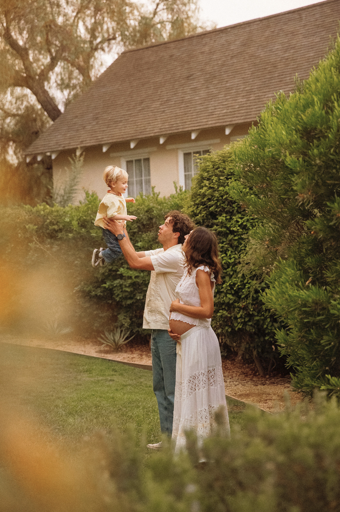 A family enjoying an outdoor moment in a garden with lush bushes and trees. A man is lifting a young boy into the air, and a pregnant woman is standing nearby, watching them.