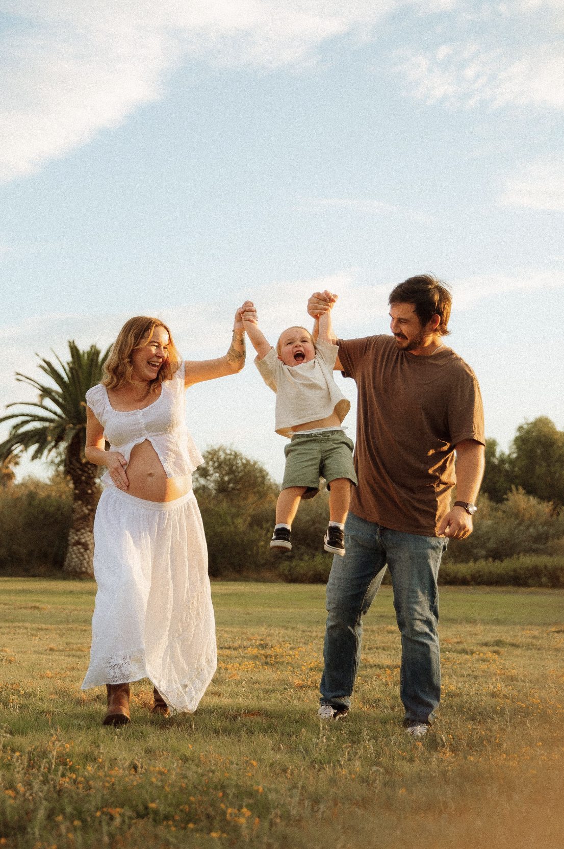 A family of three playing outside in a grassy field during sunset, with a pregnant woman, a man, and a young boy. The woman and the man are holding the boy's hands and lifting him into the air, and the boy is smiling and laughing.