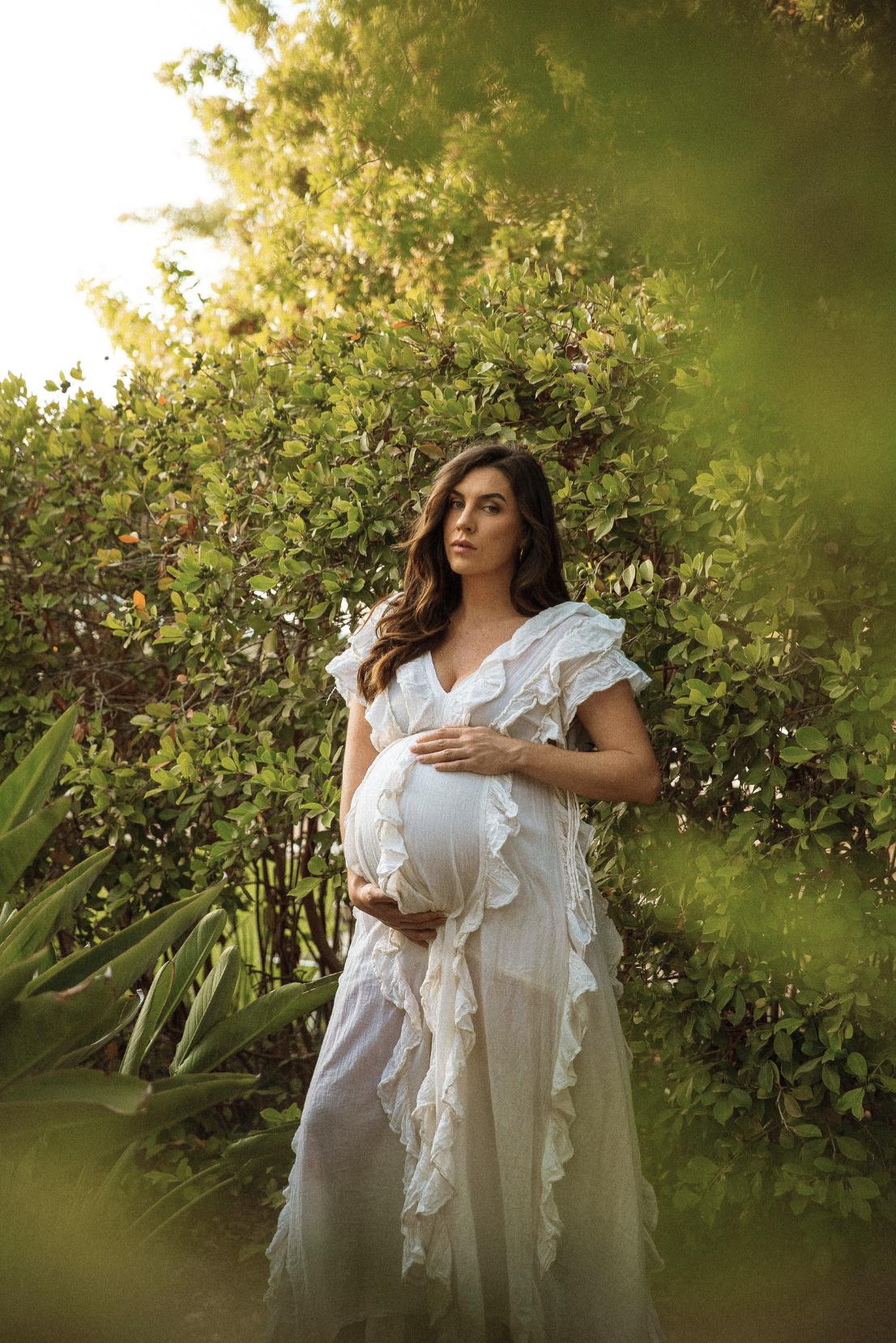 Pregnant woman in a white dress standing outdoors near green bushes, holding her belly, with sunlight filtering through trees.