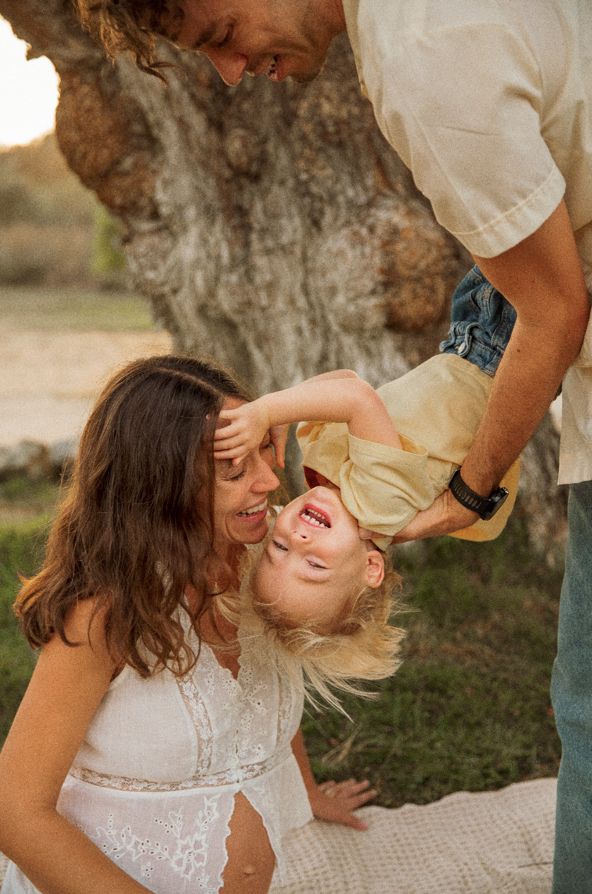 A playful outdoor scene with a woman, a man, and a young boy under a tree. The woman is sitting on the ground, smiling, while the boy is upside down, laughing and holding his head with both hands. The man is holding the boy upside down by his waist, and the background shows a natural setting with greenery and a tree.