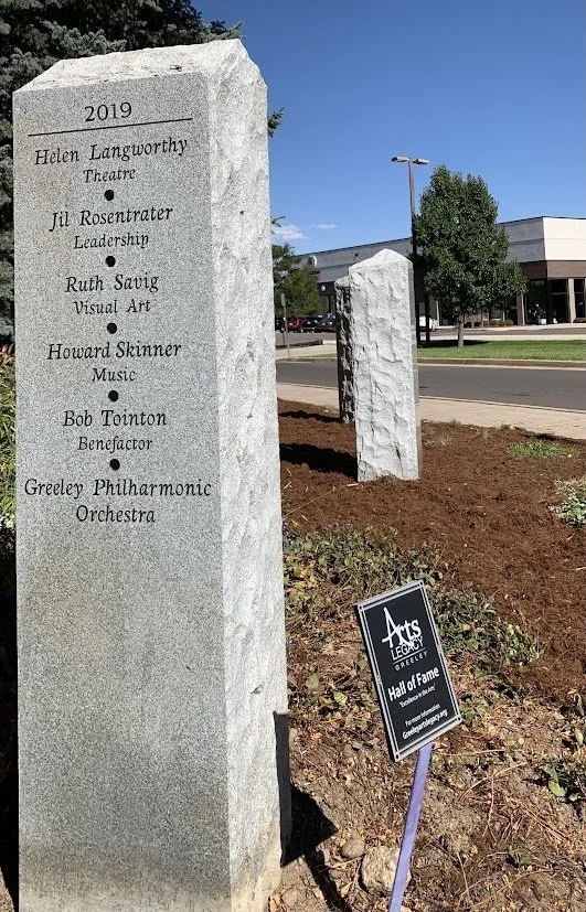 A stone monument listing names and contributions of individuals related to the arts, with a smaller black and white sign indicating that the area is part of the Greeley Arts Legacy Hall of Fame.