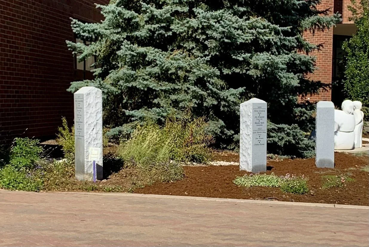 Three stone memorials are displayed in front of a large evergreen tree and a brick building.