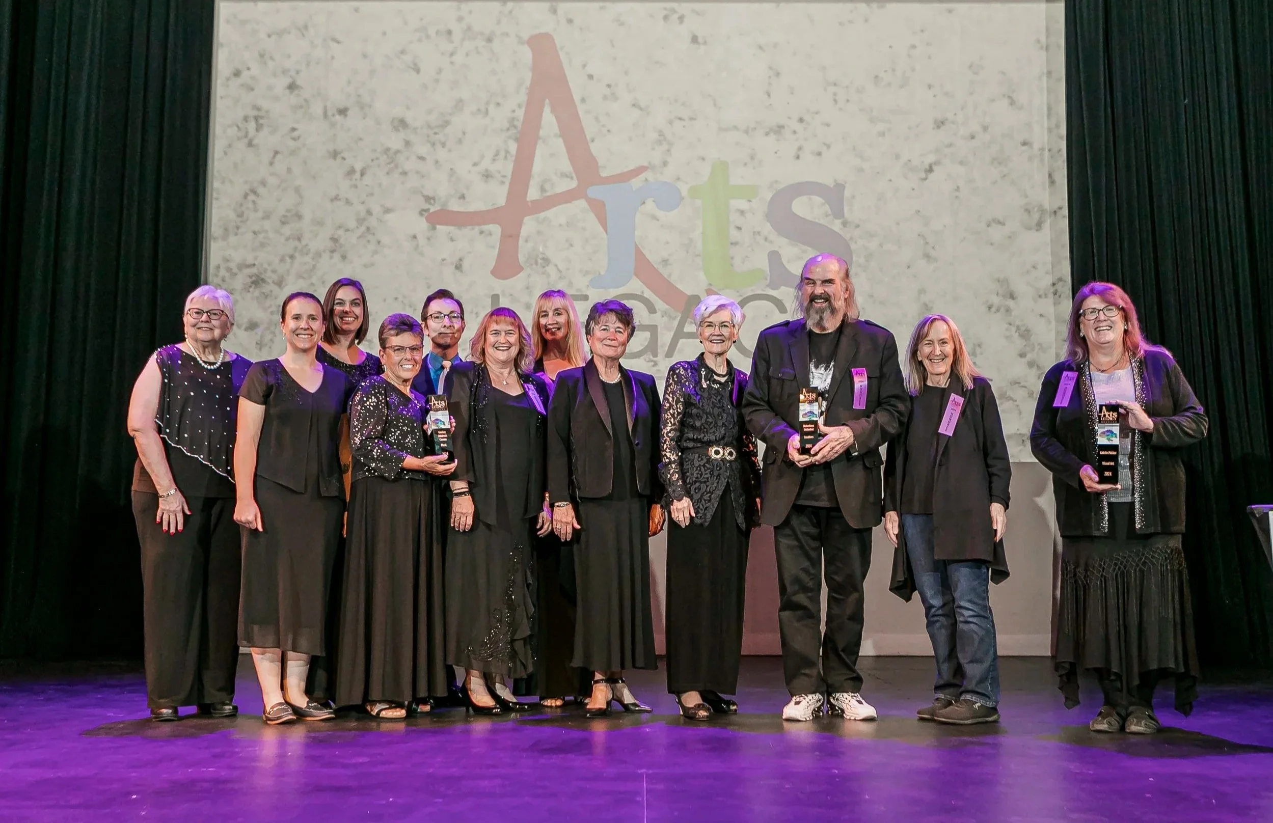A group of eleven people standing on a stage, smiling and holding awards, with a large background display reading 'Arts' and 'Gala' behind them.