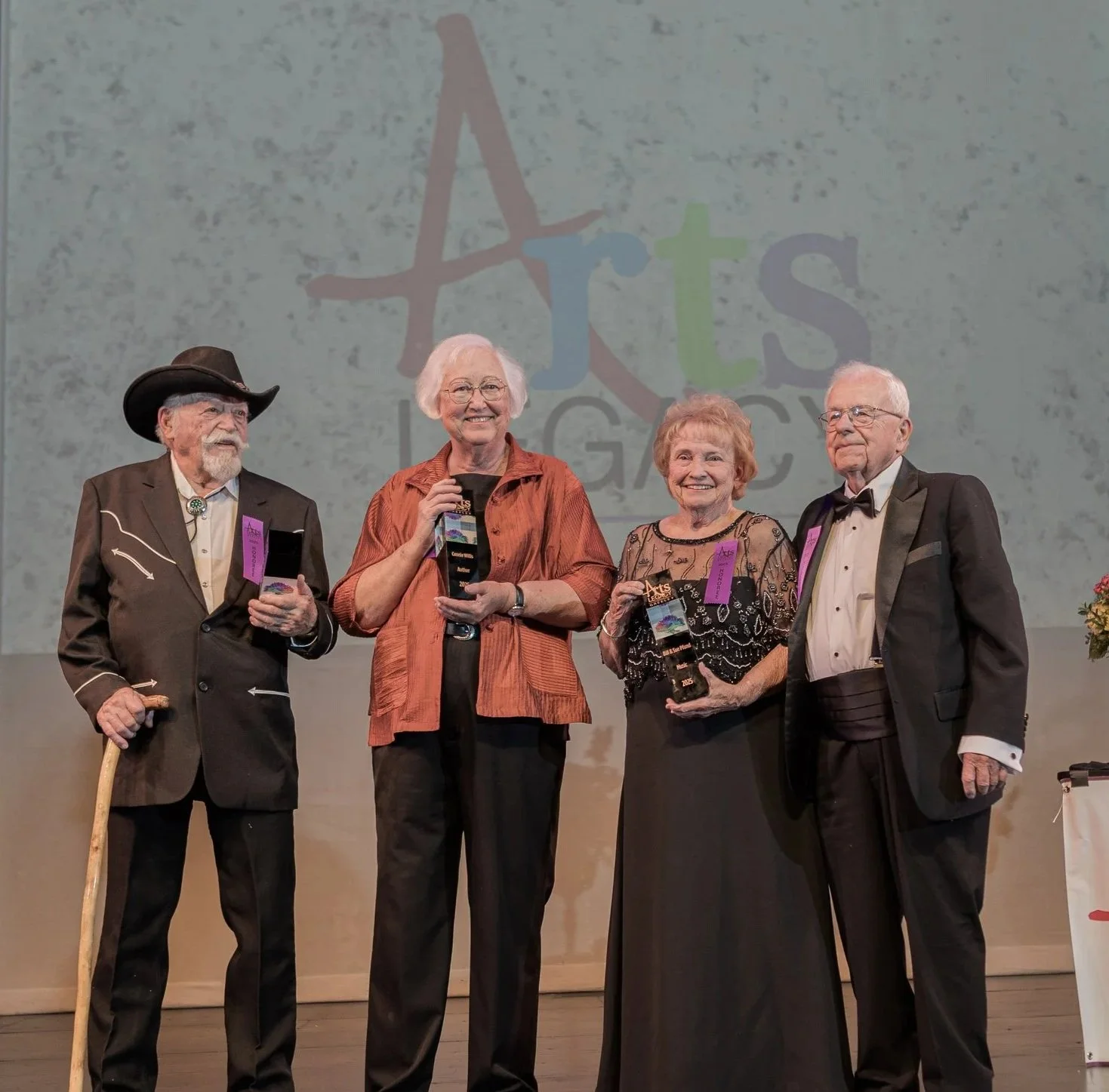 Four individuals, two men and two women, standing on a stage during an award ceremony. The women are holding trophies, and the women are smiling. One man is wearing a cowboy hat and holding a cane, while the other man is in a tuxedo. The background displays a colorful logo and the text "Arts Legacy."
