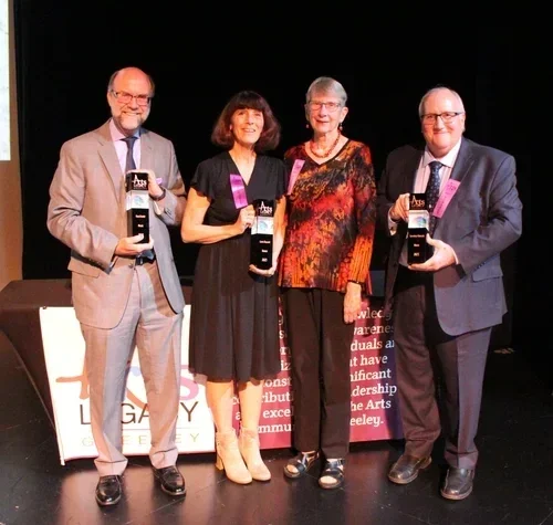 Four people standing on a stage holding awards, with a black background and a banner behind them.