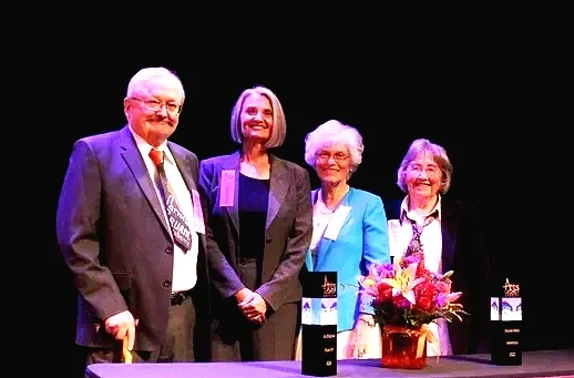 Four people standing on stage behind a table with flowers and awards, smiling.