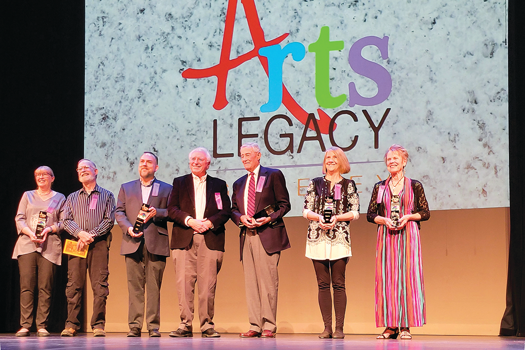 Group of seven people standing on stage holding awards at the Arts Legacy event, with a large screen displaying the event logo behind them.