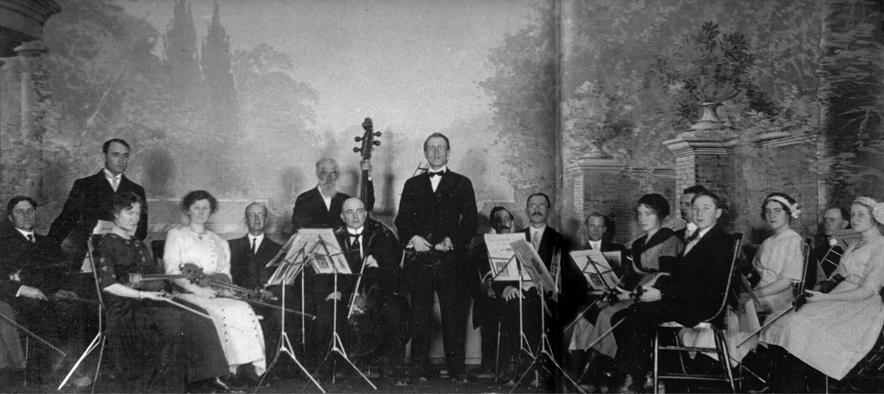 Black and white photo of a classical music orchestra with men and women in period clothing, holding instruments like violins, a cello, and a clarinet, with one standing musician playing a double bass, positioned in front of a painted backdrop of trees and columns.