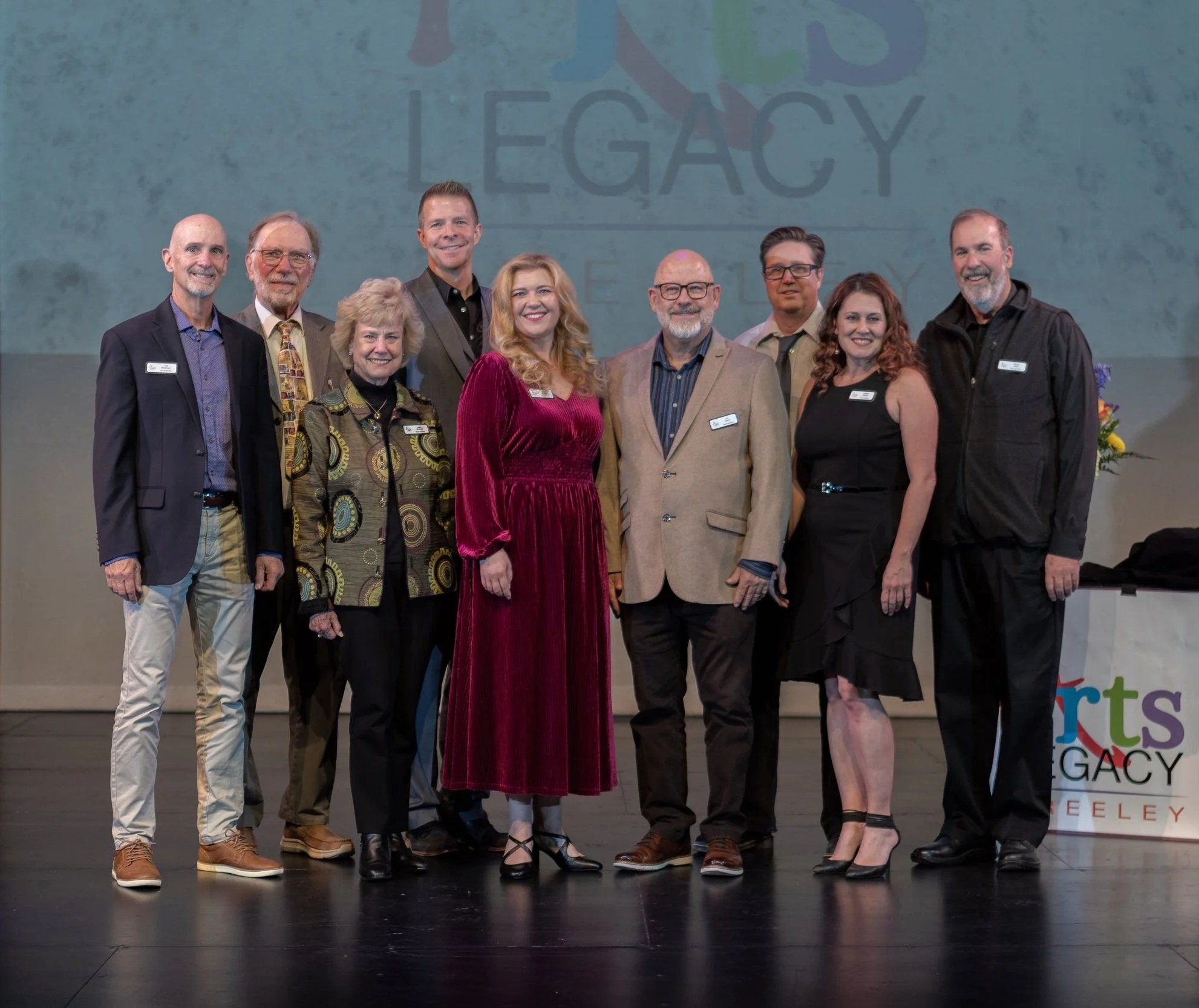 A group of nine diverse people standing on a stage during an event, with a large background screen displaying the word 'LEGACY' and colorful text. The group includes men and women dressed in professional attire, smiling for the photo.