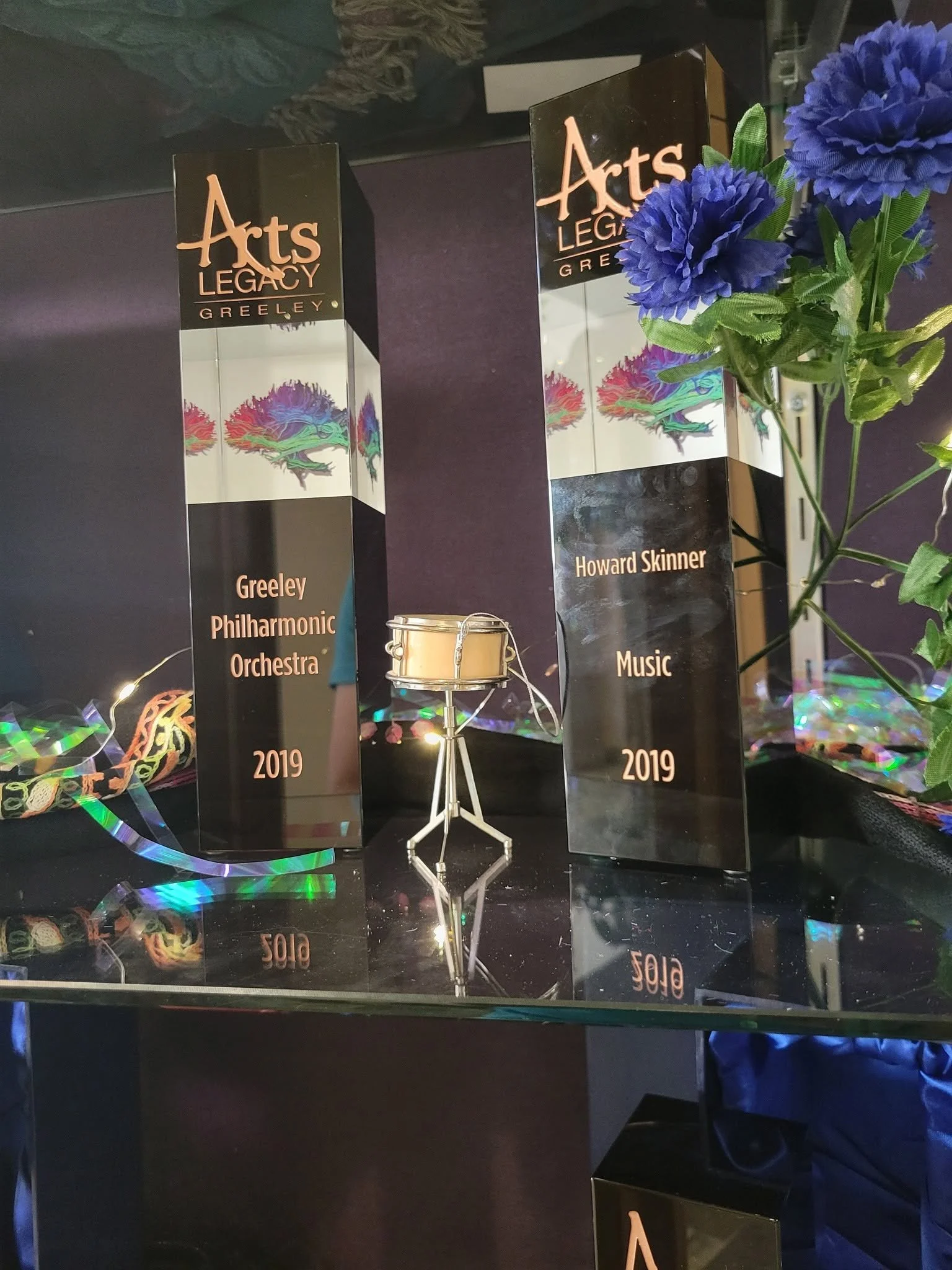 Awards plaque for Greeley Philharmonic Orchestra and Howard Skinner, with a purple flower decoration and colorful ribbons, displayed on a reflective black surface.