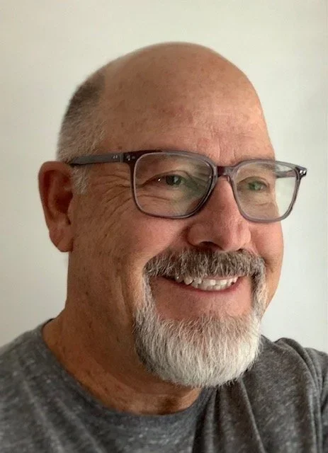 A smiling middle-aged man with glasses and a beard, wearing a gray shirt, standing against a plain light-colored background.