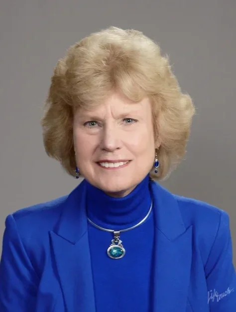 A portrait of an older woman with blonde, curly hair wearing a blue blazer, matching turtleneck, and jewelry, against a gray background.