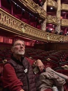 Man sitting in a theater with ornate gold and red decor, empty seats, and balconies in the background.