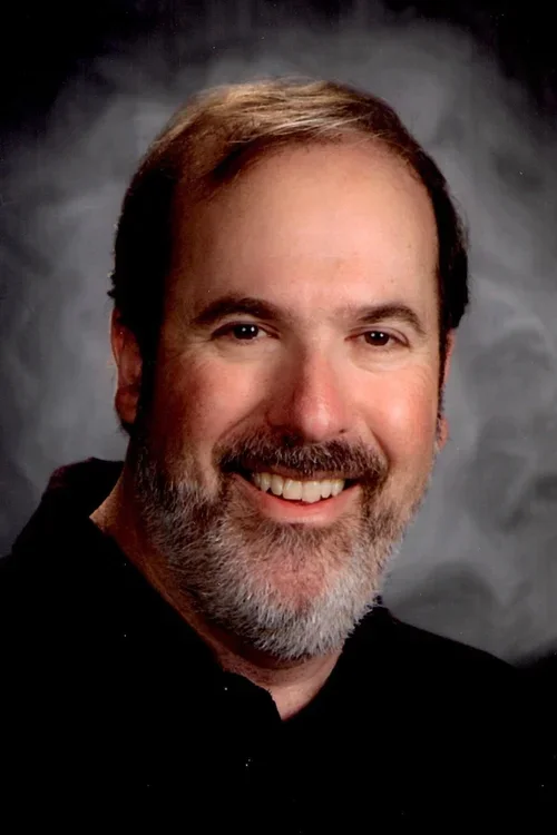 A middle-aged man with a beard and mustache, smiling in front of a gray background.