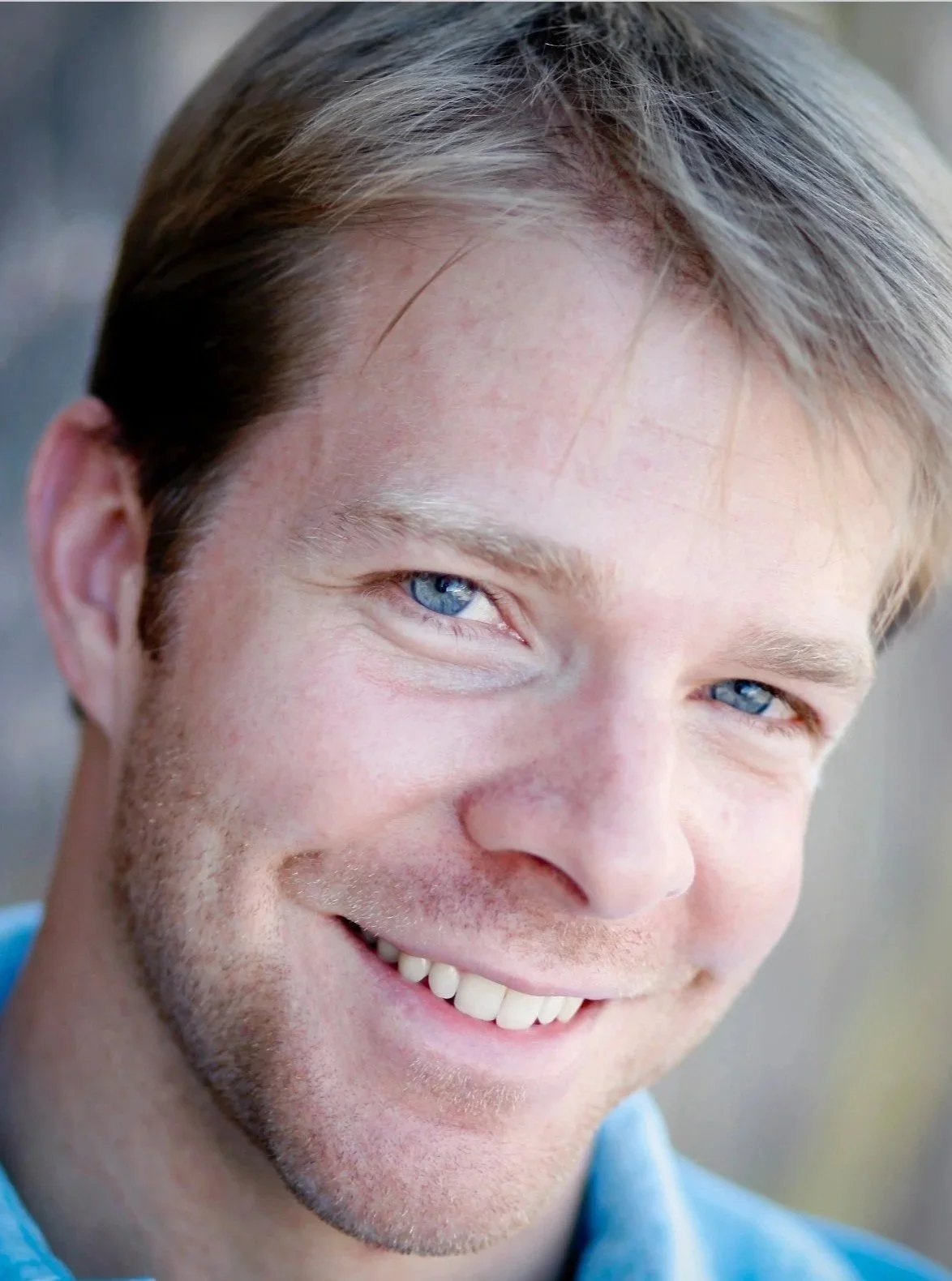 Close-up of a smiling man with blue eyes, light brown hair, and a light beard, wearing a blue shirt.