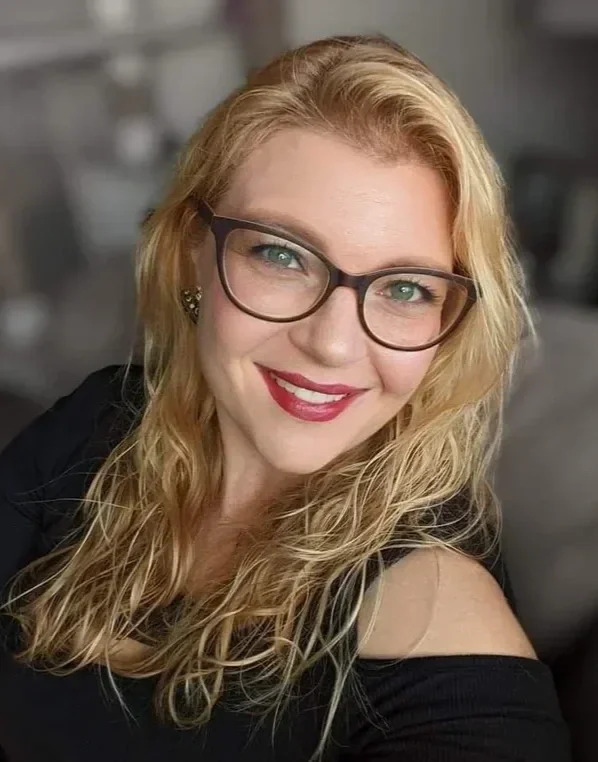 A woman with long curly blonde hair, wearing glasses with dark frames, red lipstick, and a black off-the-shoulder top, smiling at the camera.