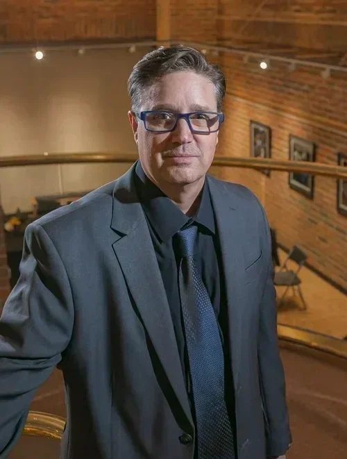 A man with glasses in a dark suit standing indoors in front of a wooden wall with framed photos.