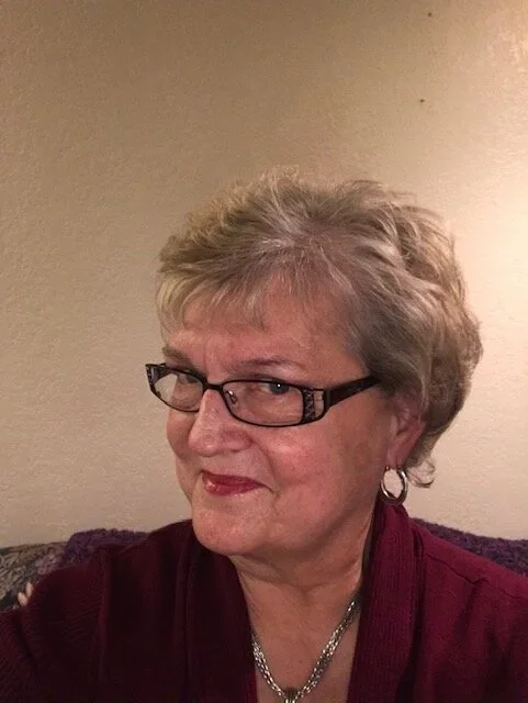 A woman with short gray hair and glasses, wearing a burgundy top, smiling slightly, sitting on a couch with a beige wall in the background.