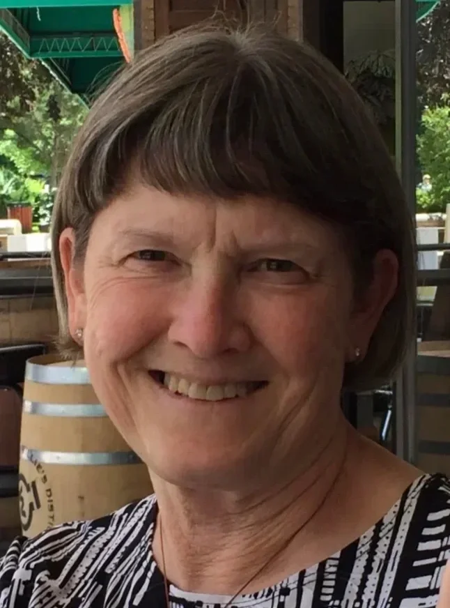 Smiling woman with short brown hair, wearing earrings, in an outdoor setting with green foliage and wine barrels in the background.