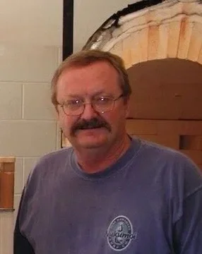 A middle-aged man with glasses and a mustache smiling, wearing a gray T-shirt, standing indoors near a stone and brick arch.