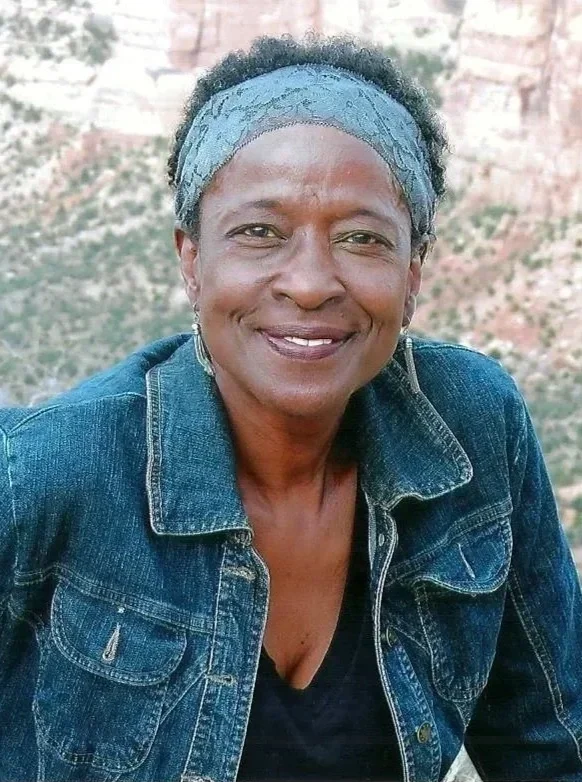 A smiling woman wearing a denim jacket and a blue headscarf outdoors with rocky landscape in the background.