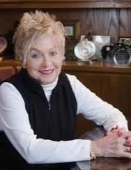A woman with short, light-colored hair smiling at the camera, sitting at a table in a room with wooden furniture and decorative items.
