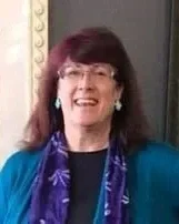 A smiling woman with dark hair and glasses, wearing a blue jacket and a purple scarf, standing indoors.