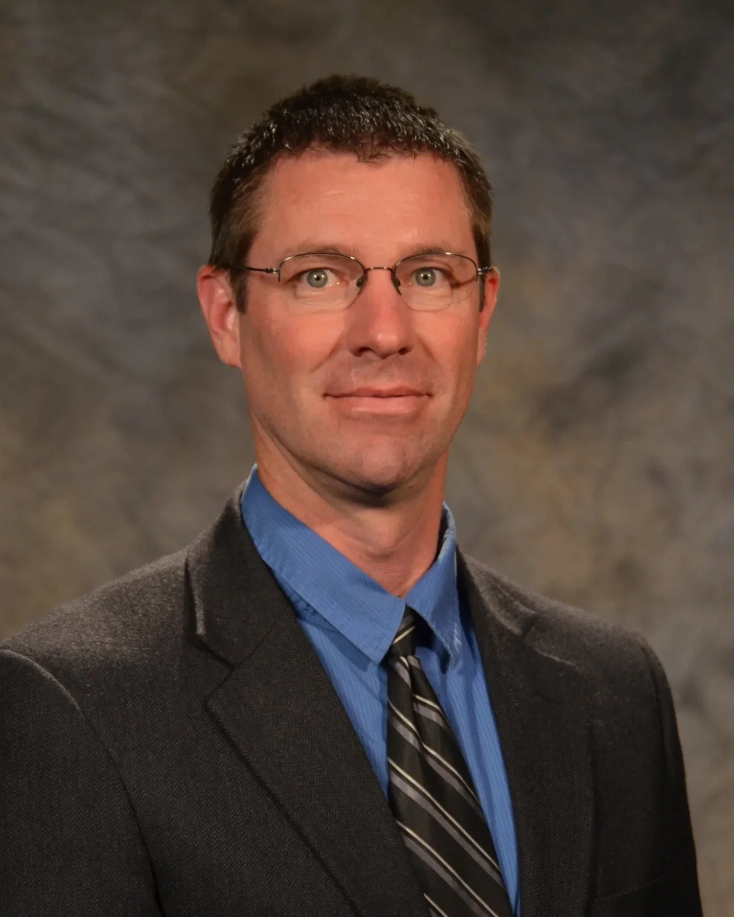 A man with short brown hair, glasses, and a slight smile, wearing a dark suit, blue shirt, and a striped tie, posing against a neutral gray background.