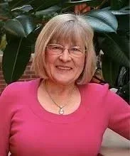 A smiling woman with shoulder-length blonde hair and glasses, wearing a pink top, standing in front of green leafy plants.