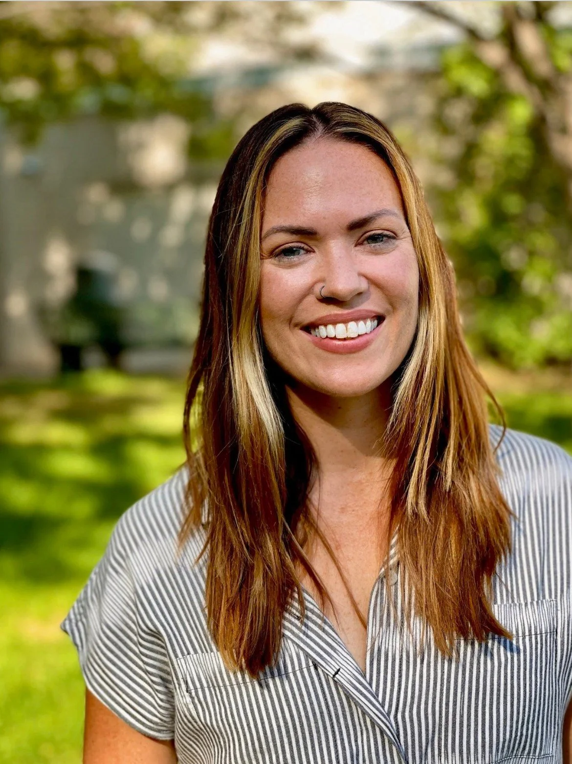 A young woman with long brown hair, smiling outdoors, wearing a striped shirt, with blurred green trees and grass in the background.
